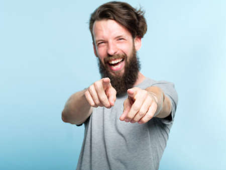 ha ha loser. young bearded hipster man mocking and laughing at viewer pointing fingers. mockery humiliation and psycological abuse concept. portrait of a grinning guy on blue background.の写真素材