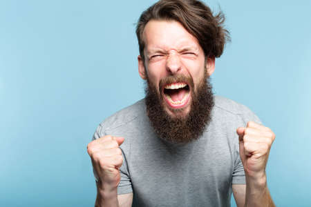 anger and fury. emotional or mental breakdown. enraged man screaming. portrait of a young bearded guy on blue background. facial expression and feelings concept.の写真素材