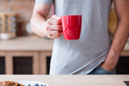 traditional morning hot beverage. quick and easy breakfast. man holding tea or instant coffee drink in a red mug standing in the kitchen.の写真素材