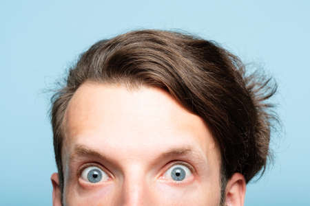 startled eccentric whimsical man with crazy look closeup. head of a young brunet guy peeking out from the bottom of blue background. emotion and facial expression concept.の写真素材