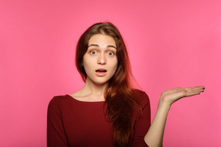emotion face. overwhelmed perplexed shocked surprised astounded woman holding virtual object on the palm of her hand. young beautiful brown haired girl portrait on pink background.の写真素材