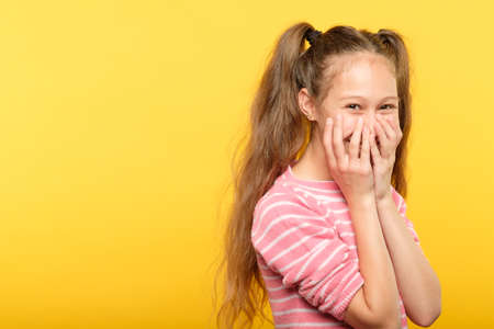 shy smiling embarrassed girl covering mouth with hands. young cute child emotional portrait on yellow background.の写真素材