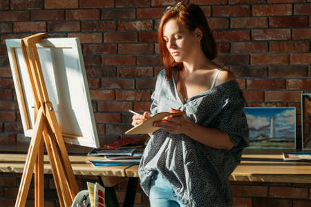 Artist painting. Studio workspace. Redhead female with easel mixing colors on wooden palette.