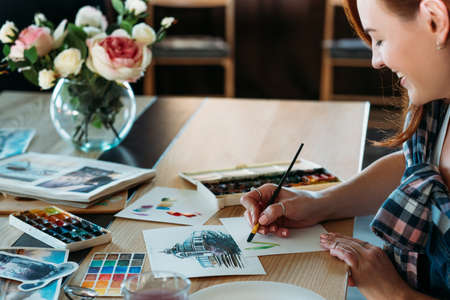 Watercolor painting. Artist at work. Smiling female painter doing color mix brushstrokes. Sketches and palette supplies around.の写真素材