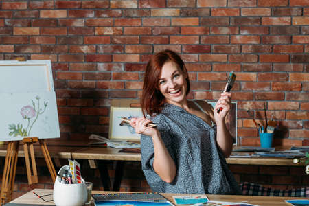 Art therapy. Stress relief studio atmosphere. Smiling redhead female posing with paintbrushes.の写真素材