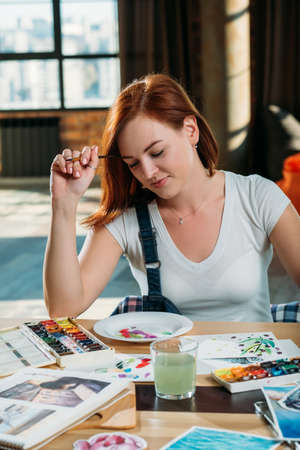 Artist workplace. Inspiration. Redhead female painter think looking at brushstrokes with sketches and palette supplies around.の写真素材