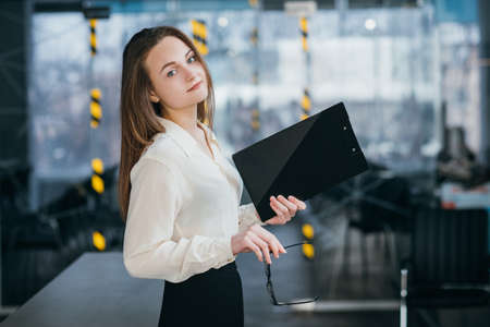 Young business woman. Successful corporate executive at workplace. Pretty female holding glasses and folder.の写真素材