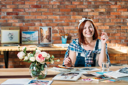 Artist painting delight. Smiling redhead female doing watercolor picture. Sketchbook and palette supplies around.の写真素材