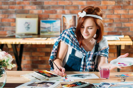 Artist at work. Watercolor painting. Redhead female painter mixing colors. Sketches and palette supplies around.の写真素材