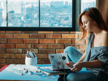 Artist inspiration. Concentration. Redhead woman painter flipping sketchbook with palette and felt pens around.の写真素材