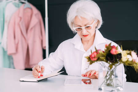 Successful senior woman. Fashion boutique business. Thoughtful elderly lady busy browsing tablet and making notes in day planner.の写真素材