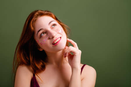 Cute young woman portrait. Choice. Redhead female looking curiously up. Copy space on green background.の写真素材