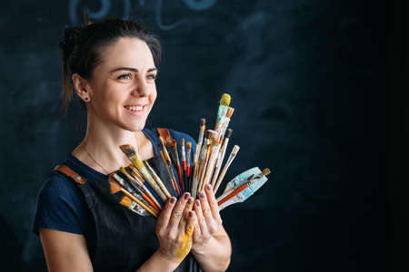 Artist supplies. Artwork in process. Smiling young woman in apron with paintbrushes assortment. Copy space for text.の写真素材
