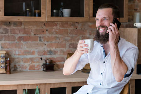 Good news at breakfast. Communication. Happy young man talking business. Latte cup and smartphone in hands.の写真素材