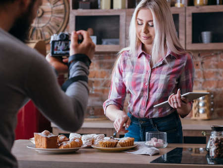 Food blogging business. Couple lifestyle. Man shooting woman pointing at muffin. Pastries assortment around.の写真素材