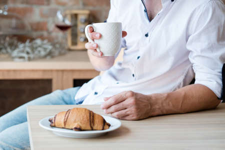 Morning coziness. Home solitude. Breakfast time. Man with cup of hot drink and croissant. Kitchen...