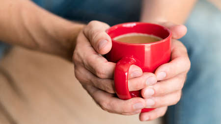 Coffee break. Relaxation and enjoyment time. Contemplation and warmth. Man hands around red cup of latte.の写真素材