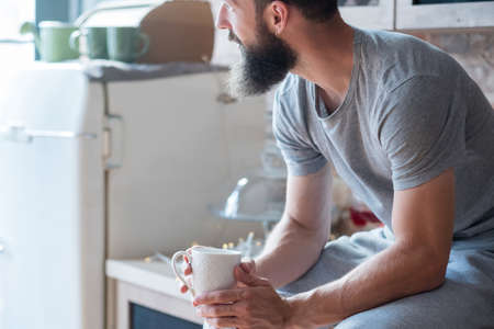 Morning coziness. Home solitude. New day. Man with cup of hot drink sitting on kitchen counter and looking sideways.の写真素材