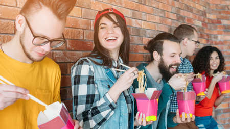 Millennials nutrition. Young coworkers having fun during lunch break, eating noodles with...