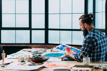 Creative working space. Young artist sitting at loft studio with big window, looking at his abstract artwork.の写真素材