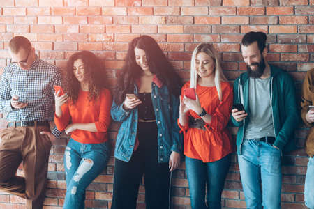 Millennial generation leisure. Diverse stylish young people leaned against wall, reading fun content on modern smartphones.の写真素材