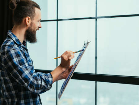 Fine art. Creative process. Handsome bearded guy inspired by view outside the window, enjoying...
