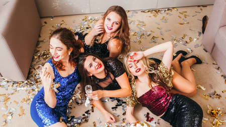 Girls party. After celebration chaos. Delighted ladies in cocktail dresses lying tired on floor with glasses, confetti around.の写真素材