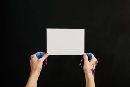 Art hobby and creativity. Painting courses. Dirty male hands holding empty paper. Invitation mockup. Dark background.の写真素材