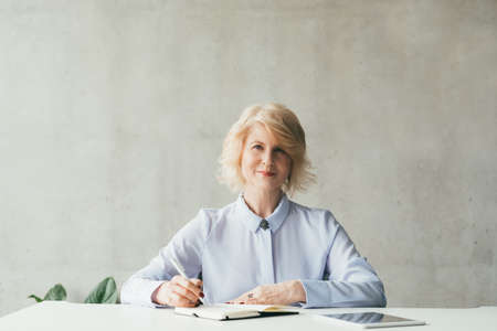 Succesful business career. Portrait of confident mature blonde woman sitting at desk with pen,...