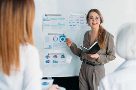 Business coaching. Successful smart woman standing at whiteboard with graphs, providing training for...