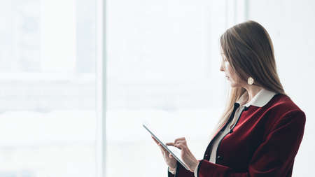Successful business lady. Corporate lifestyle. Side view of classy woman using tablet for work against big window.の写真素材