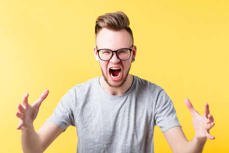 Portrait of fierce hipster guy on yellow background. Yelling young emotional man in glasses....