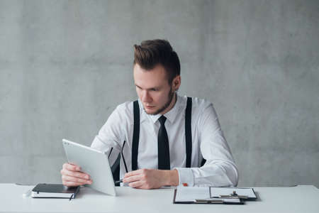 Corporate lifestyle. Portrait of young office manager sitting at desk, using tablet. Serious facial...