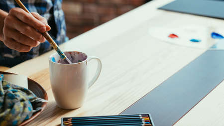 Artist workplace. Art studio atelier atmosphere. Man dipping paintbrush into white cup. Learning...