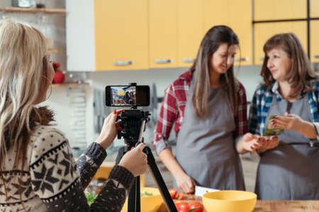 Modern culinary video blog. Family hobby. Female assistant recording video with mother and daughter cooking in kitchen.の写真素材