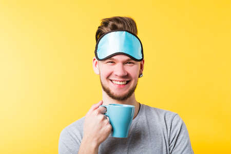 Good morning. Happy smiling young man with cup of drink beverage tea breakfast. Toothy smile sleep mask on forehead.の写真素材