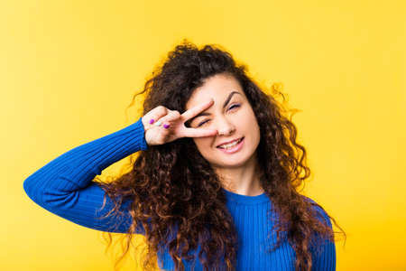 Portrait of cheerful brunette girl with curly hair. Funny emotional lady winking eye through fingers in victory gesture, smiling.の写真素材