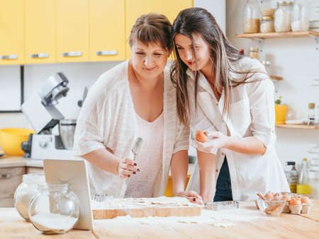 Women making pastries. Mother daughter looking at tablet. Recipe cooking class course online. Home baking hobby fun.の写真素材