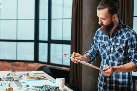 Artist studio. Man creating artwork. Board with painting in hands. Serious facial expression. Contemplation reflection.の写真素材