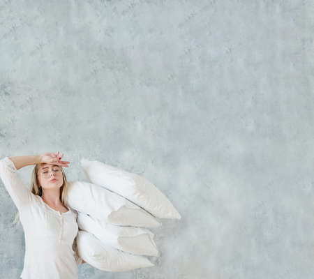 Housewife daily routine. Tired woman holding pile of pillows. Gray wall background. Copy space.