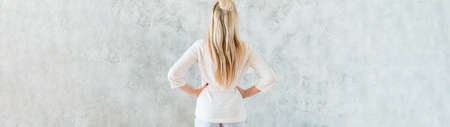 Interior design. Back view of blonde woman standing with hands on hips. Gray wall background. Copy space.の写真素材