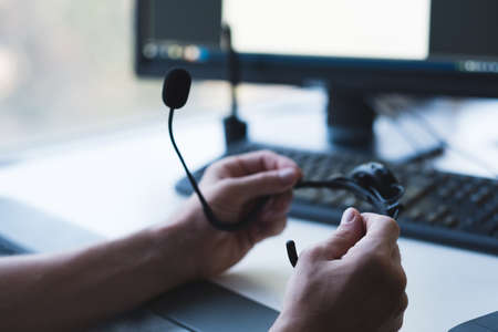 Tech support. Software engineer professional career. Closeup of headset in male hands. Blurred monitor and keyboard in background.の写真素材