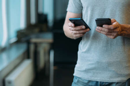 Online business communication. Cropped shot of man using two smartphones. Blur office background.の写真素材
