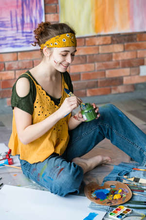Art therapy and leisure. Creative young female painter sitting on floor, washing paintbrush, creating abstract artworks.の写真素材