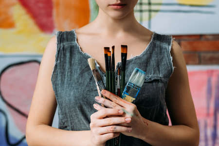 Professional artist essential tools. Cropped shot of painter holding bunch of various paintbrushes. Blur colorful background.の写真素材