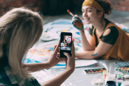 Friends leisure. Lady taking picture of young female artist posing on floor with paint and brush, smiling.の写真素材