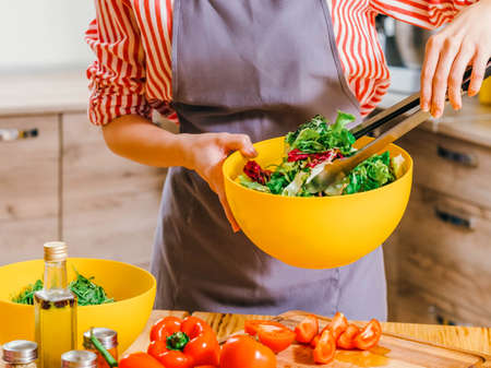 Healthy nutrition. Cropped shot of woman cooking, using kitchen tongs to mix vegetable salad in yellow bowl.の写真素材