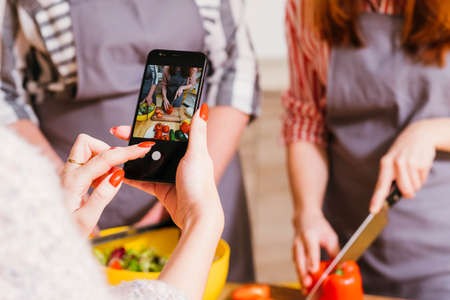 Online cooking class. Female assistant filming women preparing vegetable salad. Blur background.の写真素材