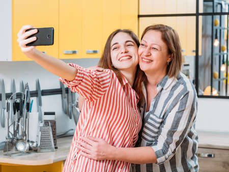 Family leisure. Mother and daughter having fun, taking selfie in home kitchen, hugging each other, smiling.の写真素材