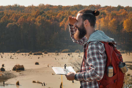 Hiking tourism. Side view of man using map and compass to explore country. Fall landscape...